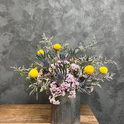 Delicate flower arrangement of yellows and purples in tall metal vase.