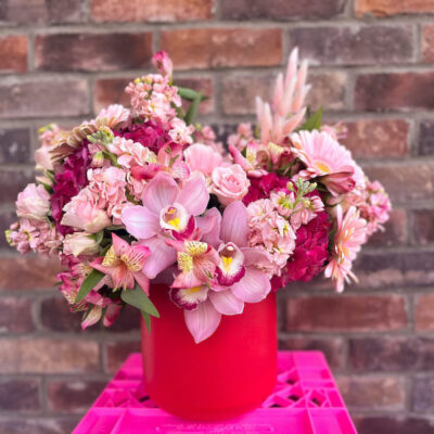 red vase filled with pink flowers sitting on a pink crate in front of a brick wall