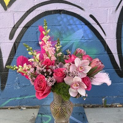 colorful flower arrangement of pink flowers and feathers in a shiny vase sitting on the ground in front of an art mural